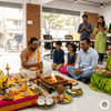 Office space mein ek Hindu puja ceremony chal rahi hai jahan ek pandit havan kar rahe hain, aas-paas parivaar aur colleagues baith kar aur khade hokar shraddha se prarthana kar rahe hain, saamne Ganesh ji ki murti aur pooja samagri sajayi hui hai.