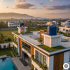An aerial, wide-angle shot of a modern villa at sunset, featuring two blue cylindrical water tanks on elevated white pedestals. The rooftop includes green turf, solar panels, and wooden pergolas, overlooking a swimming pool and distant mountains.