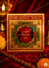 A square Shri Human Yantra plaque with intricate gold, red, and green traditional Indian designs and Sanskrit text, displayed on a red fabric altar with a brass bell and marigold flowers.