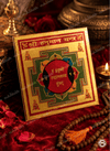 A square gold-toned Hymasan Yantra card with red, green, and black geometric designs and Devanagari text, displayed on a red velvet surface with a candle, rose petals, and wooden beads nearby.