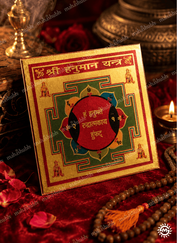 A square gold-toned Hymasan Yantra card with red, green, and black geometric designs and Devanagari text, displayed on a red velvet surface with a candle, rose petals, and wooden beads nearby.