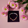 Colorful beaded bracelet on a black stand with pink flowers around it on a pink background