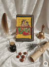 Decorative notebook with artistic cover on a textured white surface, surrounded by a feather, inkwell, bell, and rolled paper.