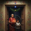 An elderly woman in a red saree and a young boy stand in a traditional Indian doorway, performing a ritual. The woman holds a lit diya (oil lamp), while the boy holds a small offering