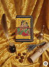 Decorative book with illustrations on a yellow fabric background, accompanied by a feather quill, small bell, and rolled paper.