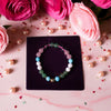 Multicolored beaded bracelet on a dark surface with pink and red flowers and pearls around it.