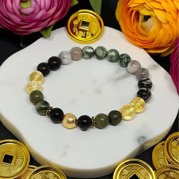 Bracelet with green and black beads and gold coins on a marble surface with flowers and more gold coins around.