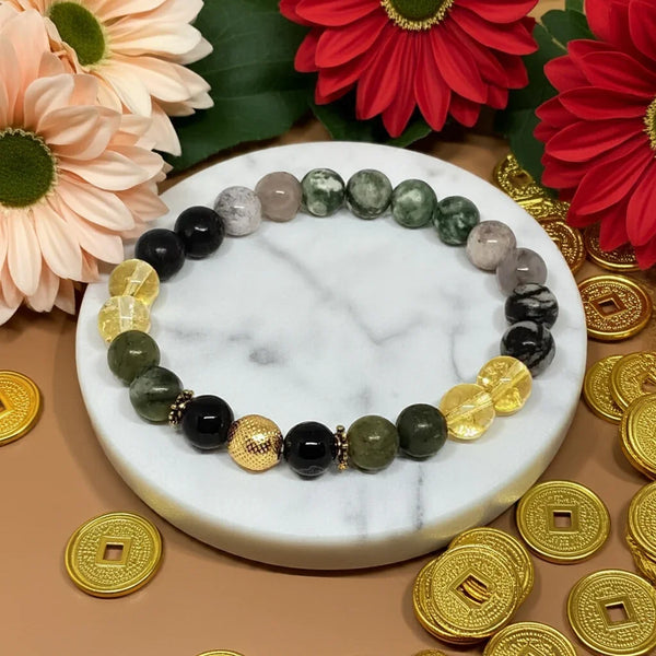 Bracelet with green, black, and gold beads on a marble surface with flowers and coins around.