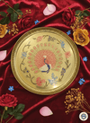 Decorative gold plate with peacock design on a red fabric background with flowers.